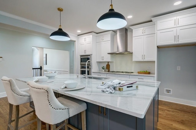 kitchen featuring stainless steel appliances, wall chimney range hood, pendant lighting, white cabinets, and an island with sink