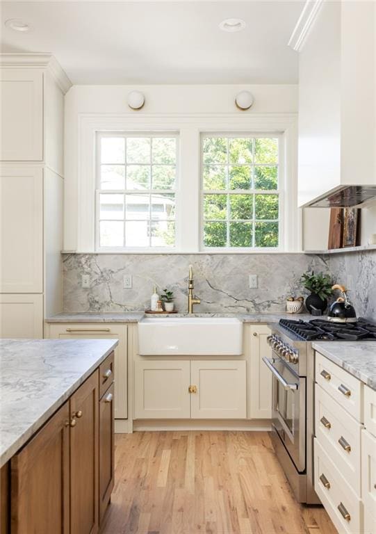 kitchen featuring light wood finished floors, light stone counters, stainless steel stove, wall chimney range hood, and a sink