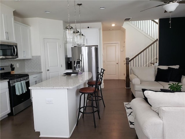 kitchen featuring dark hardwood / wood-style floors, backsplash, ceiling fan, an island with sink, and appliances with stainless steel finishes