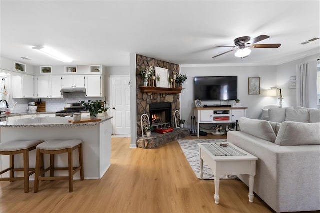 living area featuring visible vents, crown molding, ceiling fan, a stone fireplace, and light wood-style floors