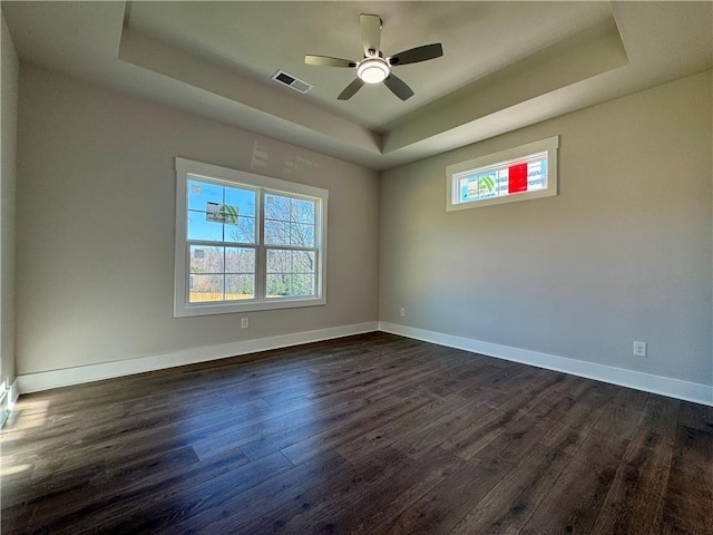 empty room with a tray ceiling, dark wood-style flooring, visible vents, plenty of natural light, and baseboards