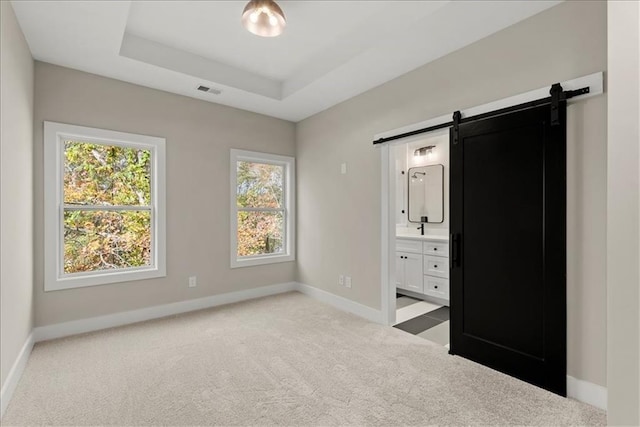 unfurnished bedroom with a raised ceiling, a barn door, ensuite bathroom, and light carpet