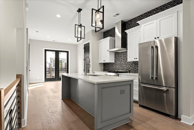 kitchen featuring wall chimney exhaust hood, stainless steel appliances, an island with sink, pendant lighting, and white cabinets