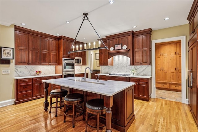 kitchen featuring light wood finished floors, an island with sink, a sink, appliances with stainless steel finishes, and tasteful backsplash