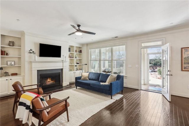 living area with a fireplace with flush hearth, built in features, baseboards, dark wood finished floors, and crown molding