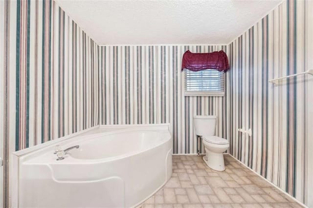 bathroom featuring a bath, a textured ceiling, toilet, and wallpapered walls