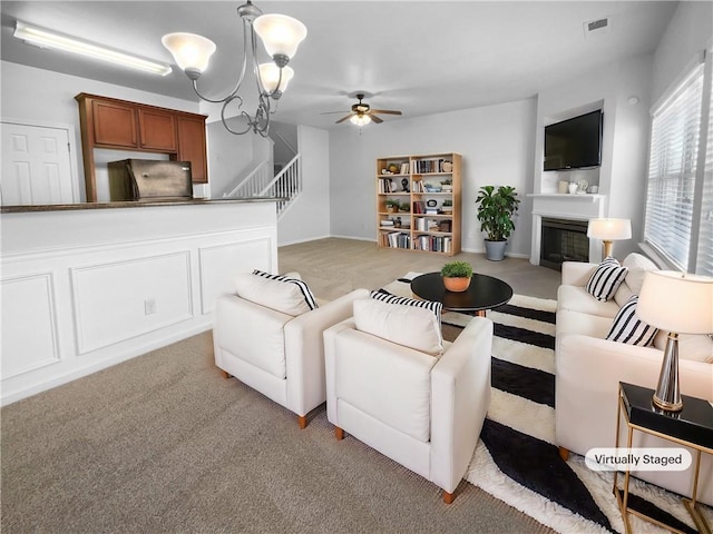 living room with visible vents, light colored carpet, a glass covered fireplace, stairs, and ceiling fan with notable chandelier