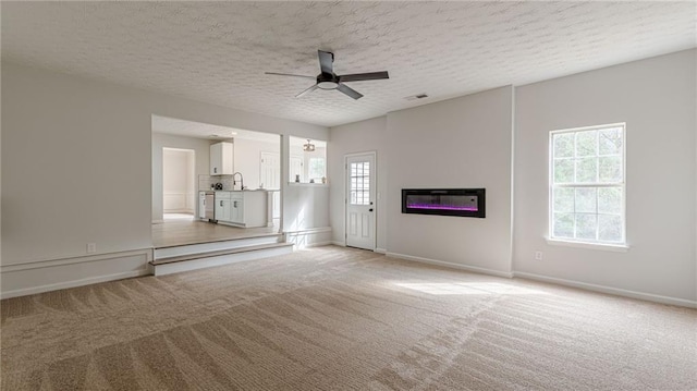 unfurnished living room featuring visible vents, light colored carpet, a ceiling fan, a glass covered fireplace, and a textured ceiling