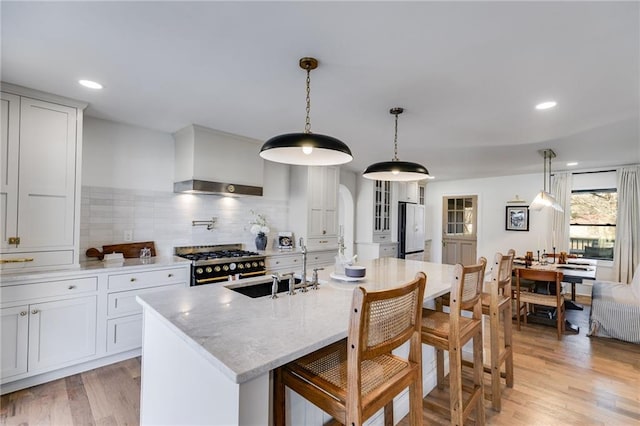 kitchen featuring pendant lighting, double oven range, a center island with sink, wall chimney exhaust hood, and white fridge