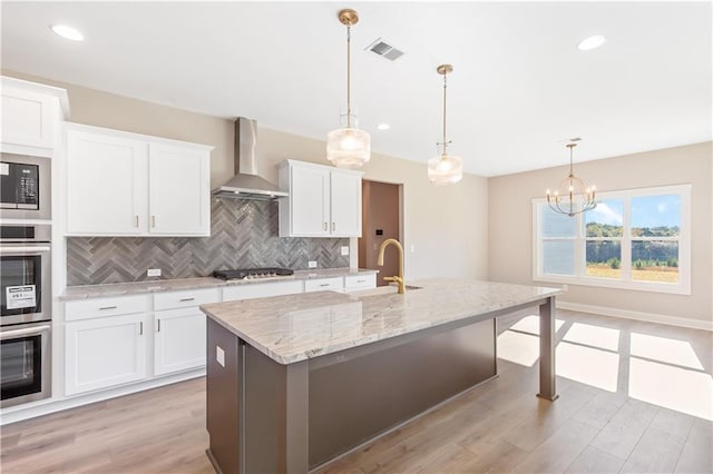 kitchen featuring white cabinetry, stainless steel appliances, wall chimney range hood, light hardwood / wood-style floors, and a center island with sink