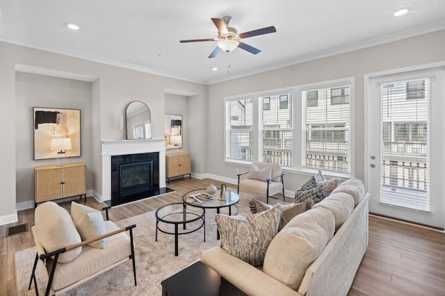 living room featuring ceiling fan, wood finished floors, crown molding, baseboards, and a glass covered fireplace