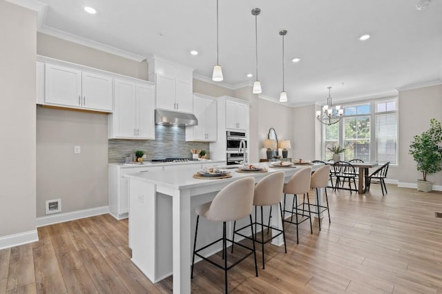kitchen with under cabinet range hood, backsplash, ornamental molding, white cabinets, and a center island with sink
