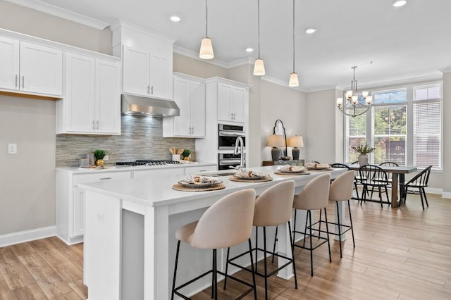 kitchen with under cabinet range hood, appliances with stainless steel finishes, an island with sink, backsplash, and light countertops