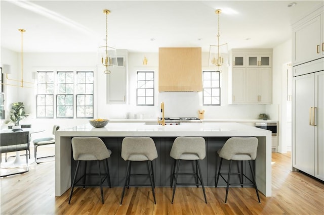 kitchen featuring white cabinets, an island with sink, hanging light fixtures, light hardwood / wood-style flooring, and paneled built in refrigerator