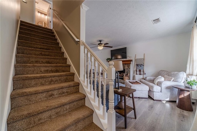 stairway with hardwood / wood-style flooring, ceiling fan, a fireplace, ornamental molding, and a textured ceiling