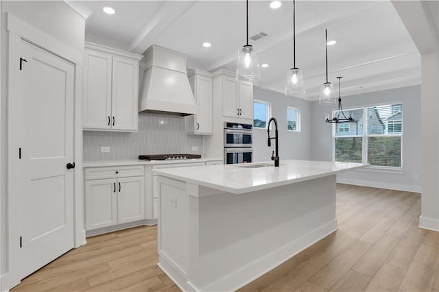 kitchen with sink, premium range hood, an island with sink, white cabinets, and decorative light fixtures