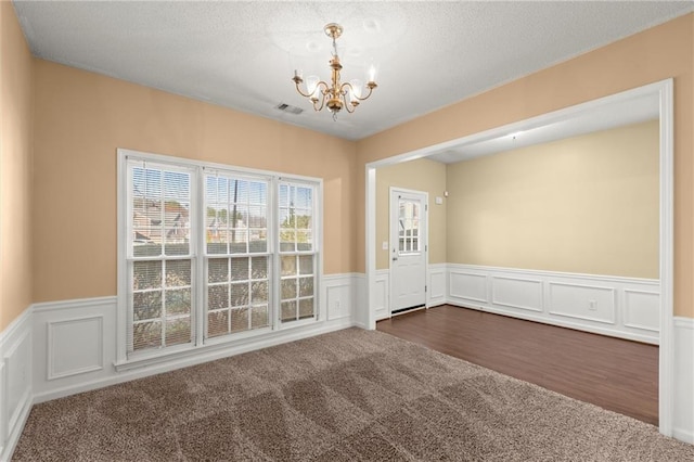 empty room featuring visible vents, a wainscoted wall, a notable chandelier, a textured ceiling, and dark carpet