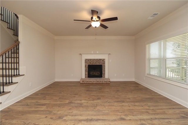 unfurnished living room featuring stairway, wood finished floors, ceiling fan, ornamental molding, and a brick fireplace