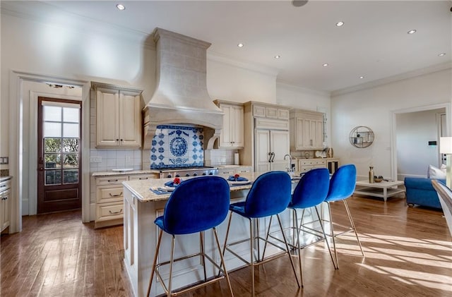 kitchen with tasteful backsplash, an island with sink, premium range hood, cream cabinetry, and hardwood / wood-style flooring