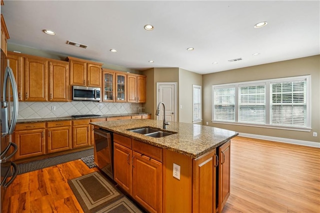kitchen with stainless steel appliances, light wood-style flooring, a sink, a center island with sink, and brown cabinets