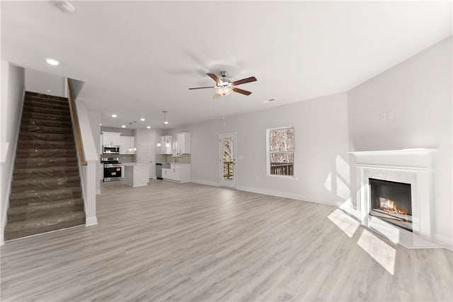 unfurnished living room with stairway, light wood-type flooring, baseboards, ceiling fan, and a fireplace