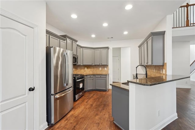 kitchen featuring dark wood-style floors, gray cabinets, appliances with stainless steel finishes, dark stone countertops, and a peninsula