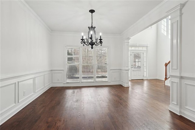 unfurnished dining area with a notable chandelier, decorative columns, crown molding, a decorative wall, and dark wood-type flooring