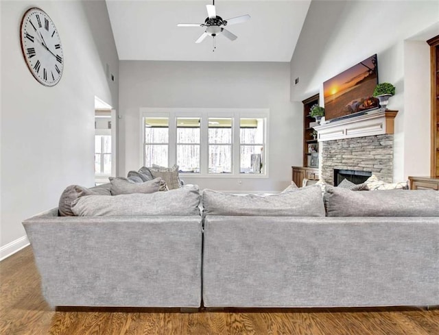 living area featuring a ceiling fan, baseboards, a stone fireplace, and dark wood-type flooring