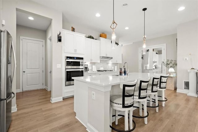 kitchen with decorative light fixtures, an island with sink, stainless steel appliances, and light hardwood / wood-style floors