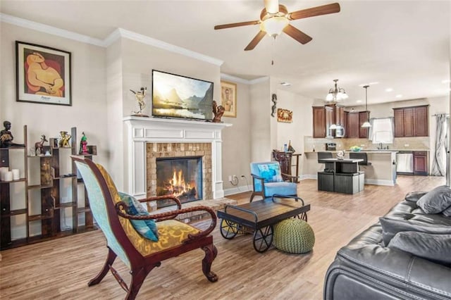 living room with a brick fireplace, ornamental molding, ceiling fan, sink, and light hardwood / wood-style floors