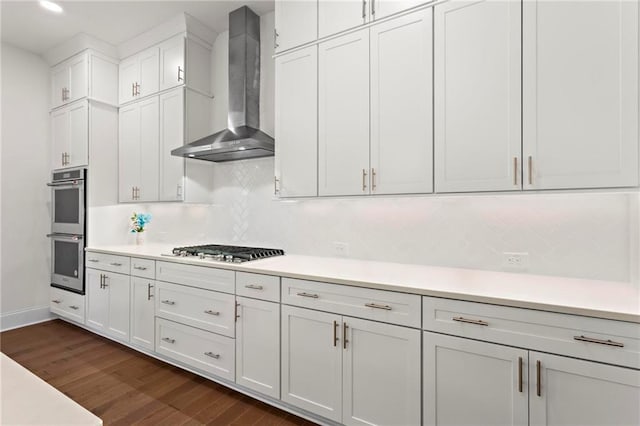 kitchen with white cabinets, wall chimney exhaust hood, light countertops, appliances with stainless steel finishes, and dark wood-type flooring