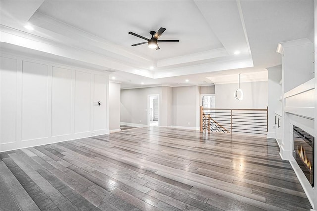 unfurnished living room featuring a glass covered fireplace, wood finished floors, a tray ceiling, crown molding, and a decorative wall