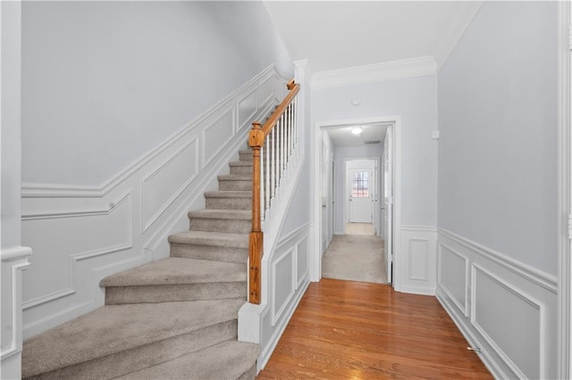staircase with wood finished floors, a wainscoted wall, crown molding, and a decorative wall
