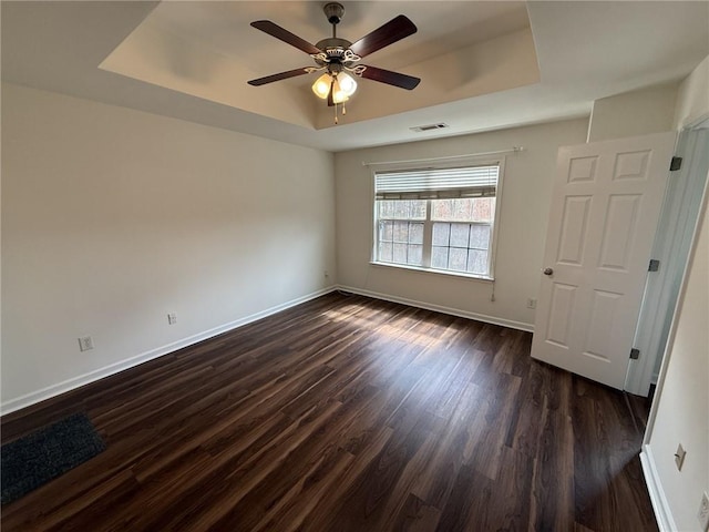 empty room with visible vents, a ceiling fan, a tray ceiling, baseboards, and dark wood-style flooring