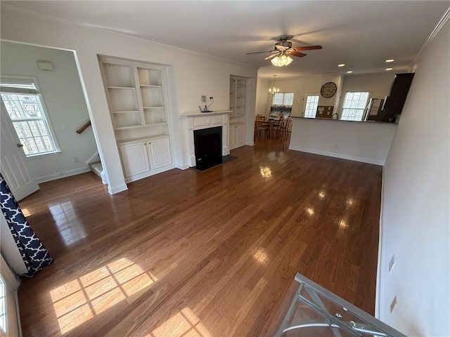 unfurnished living room featuring built in features, wood finished floors, baseboards, a fireplace with flush hearth, and crown molding