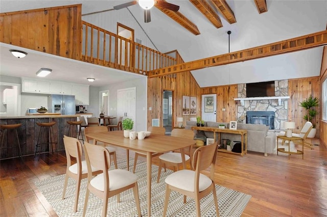 dining space featuring a stone fireplace, wood-type flooring, high vaulted ceiling, beam ceiling, and wood walls