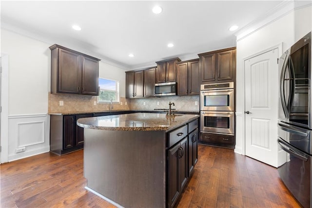 kitchen with stainless steel appliances, a kitchen island with sink, dark brown cabinetry, and dark stone counters