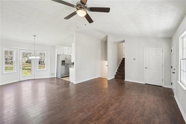 unfurnished living room with dark wood-style floors, stairway, ceiling fan with notable chandelier, and baseboards