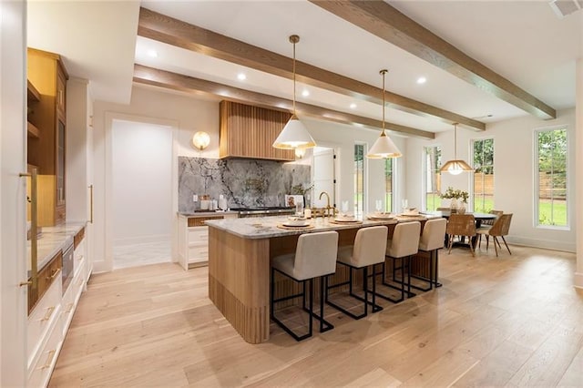 kitchen with pendant lighting, backsplash, light hardwood / wood-style flooring, light stone countertops, and an island with sink