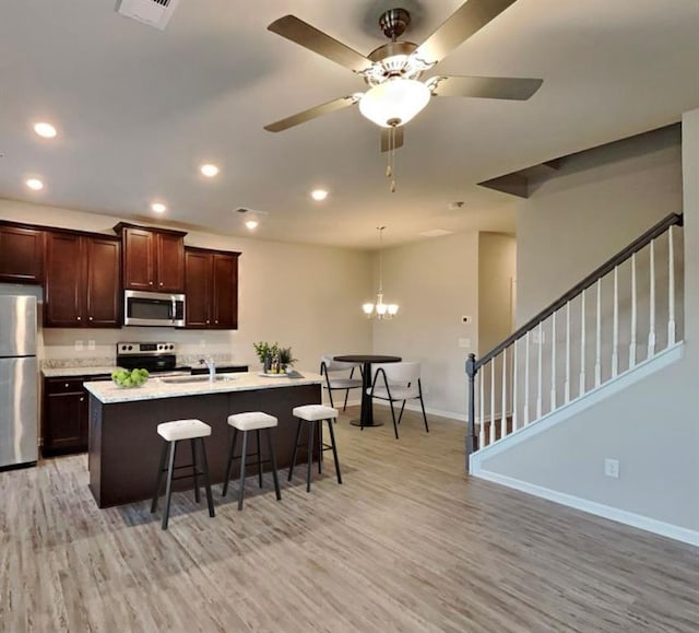 kitchen featuring a kitchen island with sink, ceiling fan with notable chandelier, light wood-type flooring, appliances with stainless steel finishes, and a kitchen bar