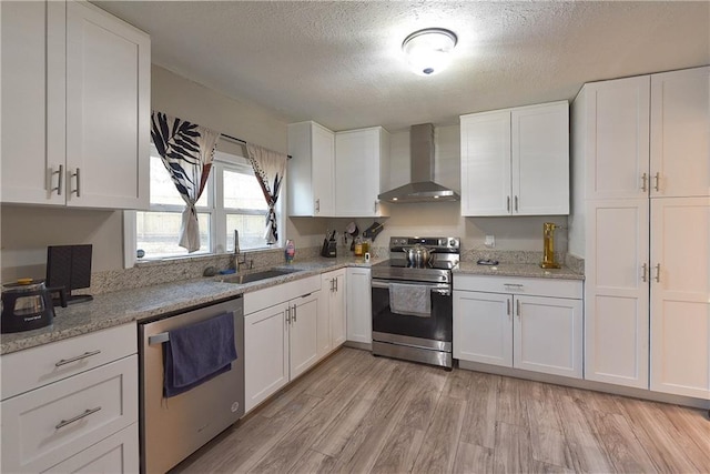 kitchen featuring light wood finished floors, appliances with stainless steel finishes, white cabinets, a sink, and wall chimney exhaust hood