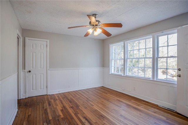 empty room with a textured ceiling, ceiling fan, a wainscoted wall, visible vents, and wood-type flooring