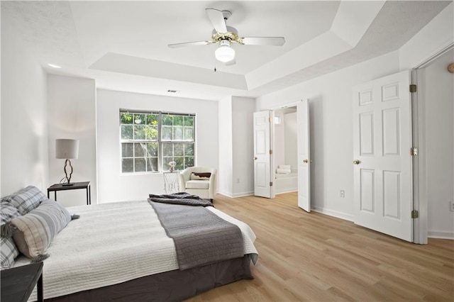 bedroom with light wood-type flooring, a tray ceiling, connected bathroom, baseboards, and ceiling fan