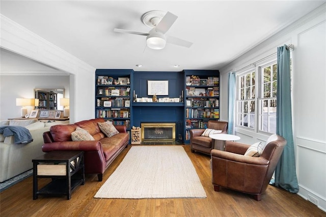 living area featuring a glass covered fireplace, a ceiling fan, wood finished floors, and crown molding