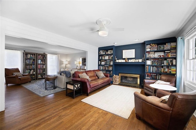 living room with built in shelves, wood finished floors, a fireplace with flush hearth, ceiling fan, and ornamental molding