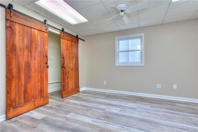 unfurnished bedroom featuring ceiling fan, a barn door, a drop ceiling, and light hardwood / wood-style flooring