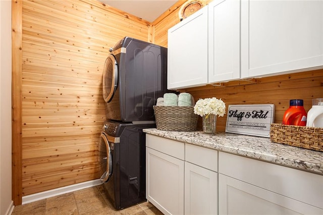 laundry area featuring cabinet space, stacked washer / drying machine, wood walls, and baseboards