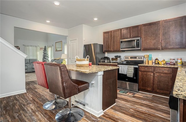 kitchen featuring light stone counters, an island with sink, a kitchen bar, dark hardwood / wood-style flooring, and appliances with stainless steel finishes