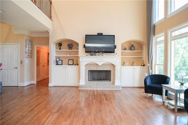 living room featuring crown molding, built in features, a high ceiling, a tiled fireplace, and light wood-type flooring