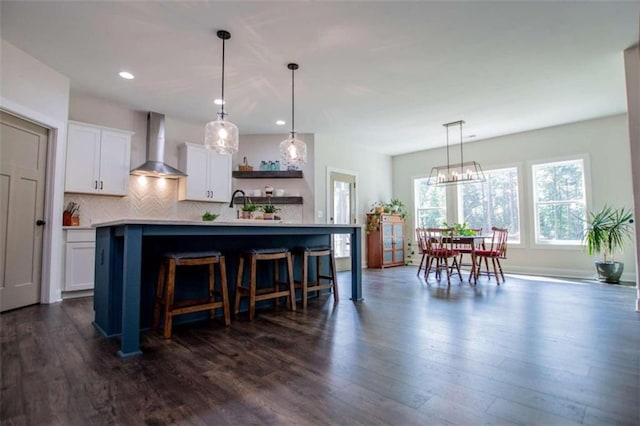 kitchen with dark hardwood / wood-style floors, wall chimney exhaust hood, pendant lighting, a kitchen island with sink, and white cabinets